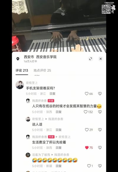 图片展示了西安音乐学院的一个场景，有人正在弹钢琴，但画面中最引人注目的是放在钢琴上的手机。下方是该地点的评论区，其中一条评论"手机支架很难买吗?"引发了有趣的对话。作者回复"人只有在低谷的时候才会发掘其智慧的力量"，被要求"说人话"后坦白"生活费没了所以先咬着"，幽默地解释了为什么用嘴咬着手机代替支架。