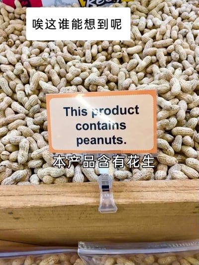 这张图片呈现了一处售卖带壳花生的场景，大量带壳花生堆积在容器中，在花生堆前方立着一块提示牌，牌面用英文标注“This product contains peanuts.”，下方配有对应的中文翻译“本产品含有花生”，图片顶部还添加了吐槽文字“唉这谁能想到呢”。该提示牌的内容完全是显而易见的，属于形式化且毫无必要的产品提示，整体内容带有强烈的搞笑与讽刺意味，调侃了这种过度刻板、脱离实际的规范执行行为。