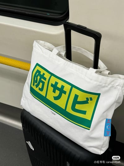 这是在地铁车厢内拍摄的出行场景，一只印有绿黄配色日文「防サビ」（意为防锈）的白色帆布袋，套在黑色行李箱的拉杆上，袋子设计参考了日本公共标识风格，简约又带有日系趣味，很适合出行时用来分装物品，和行李箱的搭配也很方便。图中没有双关、谐音梗或暗示内容。