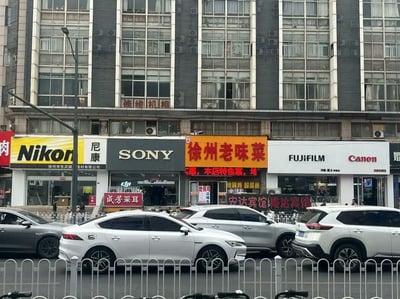 这张街头照片拍摄自徐州某街区，展现了一排连锁品牌门店，包括Nikon（尼康）、SONY、FUJIFILM、Canon等知名相机品牌。但在这几家高科技门店之间，突兀地夹着一家“徐州老味菜”餐馆，店招用鲜明的红黄配色，与左右科技风格的门头形成极强反差。这种混搭带来强烈违和感和幽默效果，令人产生“餐饮也成相机大牌之一”的错觉。视觉上很魔性，非常容易引发社交平台二次创作或调侃。