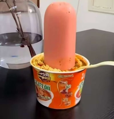 这张图通过物品摆放的位置制造视觉错位效果，将一根放在杯面后方的火腿肠，看起来像是直接插在杯面当中，营造出超大火腿肠搭配方便面的夸张搞笑画面，场景设置在普通桌面，旁边还有一台饮水机，利用视角差带来趣味反差感。
**视觉错位说明**：实际火腿肠并未插入杯面，只是在杯面后方的位置，通过拍摄角度让两者看起来重叠，形成错觉。