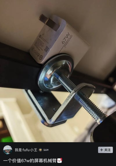 整张图为社交媒体内容截图，上方实拍部分显示屏幕机械臂的金属C型夹具安装在桌面边缘时，恰好将一个标注有「67W」字样的白色快充充电头挤压在夹具顶端与桌面底部之间，充电头被夹具牢牢压住；下方是内容发布者信息栏，显示发布者昵称为「我是fufu小王」、等级为Lv.21，配文为「一个价值67w的屏幕机械臂」搭配搞怪表情，利用功率单位「W」和中文计数单位「万」的谐音制造笑点，也对应原标题吐槽充电头被挤压的状态。