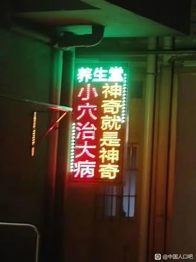 这是一张夜晚拍摄的线下养生堂的竖版LED宣传灯牌，灯牌以三色灯光呈现标语：顶部绿色字体写着“养生堂”，左侧红色字体为“小穴治大病”，右侧黄色字体是“神奇就是神奇”。灯牌旁的金属杆与灯光恰好形成类似箭头的视觉效果，指向“小穴治大病”的字样，意外增添了趣味感。该灯牌是养生店铺的宣传内容，主打穴位疗法可治疗重病的夸张宣传点，没有双关或谐音类的梗。