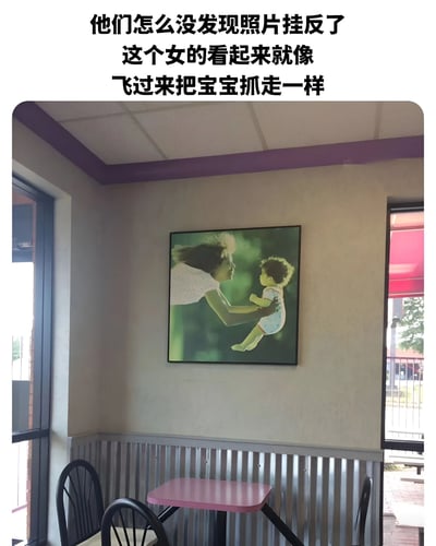图片展示了一家餐厅的角落场景，墙角挂着一幅明显倒挂的人物照片。照片中原本应是女子温柔抱起婴儿的温馨画面，但因悬挂方向错误，从观者角度看，女子头发上扬、身体前倾，姿态酷似正飞扑过来抓取婴儿，形成滑稽的视觉效果。图片上方配有文字点评这一乌龙：“他们怎么没发现照片挂反了 这个女的看起来就像飞过来把宝宝抓走一样”，突出了倒挂照片带来的搞笑误解。