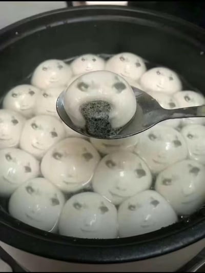 这是一张经过图像编辑软件二次创作的搞笑图片，将煮在锅中的芝麻馅汤圆表面都合成了同款真人笑脸，其中一个汤圆被勺子舀起，黑芝麻馅正从汤圆的“脸部”位置流出，原本普通的美食场景因为人脸的加入变得荒诞又滑稽，充满整活趣味，没有双关或谐音梗。