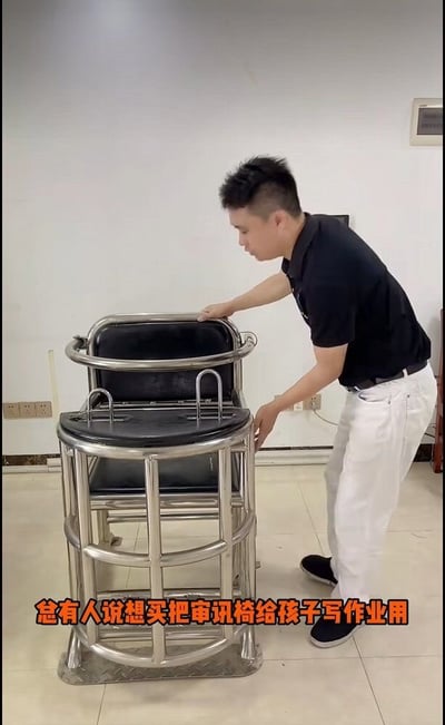 整张图为视频截出的静态实拍画面，室内场景中一名身着黑色短袖Polo衫、白色长裤的短发男子站在一把全金属不锈钢材质的审讯椅旁，双手扶着审讯椅的边缘低头查看。这把审讯椅带有约束用的扶手、卡扣结构，整体设计坚固，具备限制坐者行动的功能。画面底部配有醒目的橙色加粗字幕“总有人说想买把审讯椅给孩子写作业用”，内容是调侃部分家长为了让孩子乖乖写作业想要使用约束性工具的想法，后续也衍生出了其他方向的调侃内容。