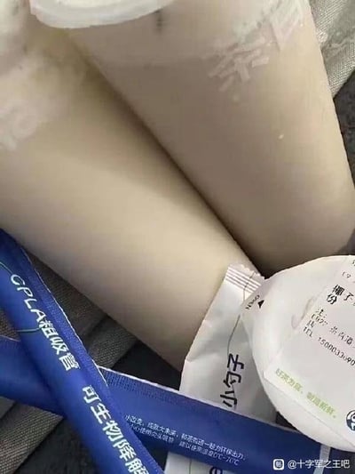 图片特写展示了多个浅色圆柱形奶茶杯堆叠在一起，杯身印有模糊的文字。旁边散落着蓝色包装的“CPLA 可降解吸管”和白色的“小勺”包装袋，吸管包装上还标注了原料及环保相关说明。整体为日常饮品周边物品的近距离拍摄，画面聚焦于奶茶容器与配套环保吸管，无明显双关、谐音梗或暗示内容。