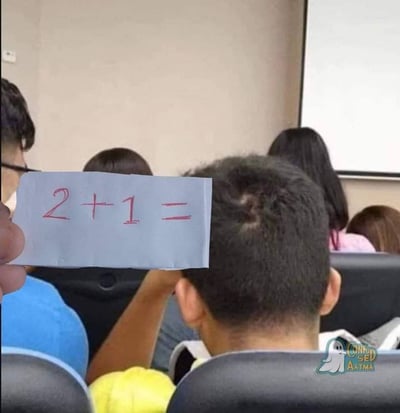 这张图拍摄于室内课堂或培训会场景，前景左侧有一只手举着一张写有红色笔迹算术题“2+1=”的浅蓝色便签，便签位置刚好对准前方穿黄色上衣男生的后脑勺，该男生头顶正好有三个发旋，通过视觉错位，三个发旋刚好承接在等号之后，组成了完整的等式“2+1=3”，创意十足充满趣味，背景还能看到其他参会或上课人员的背影、墙面以及投影幕布，右下角带有相关标识水印。