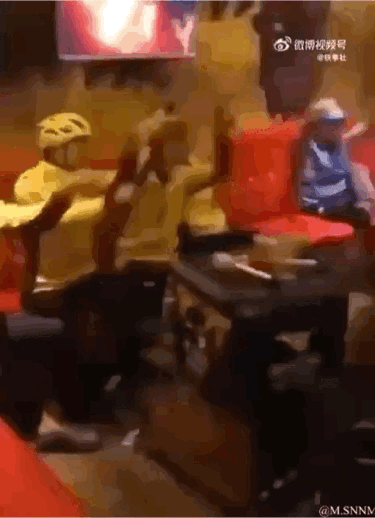这是一段动态GIF，画面场景为餐馆内部，两位分别身着黄色、蓝色外卖制服并佩戴头盔的外卖员，坐在餐桌旁的椅子上，做出幅度夸张的肢体动作，看起来像是在玩闹互动，充满了生活化的欢乐感，展现了外卖员在工作间隙的轻松时刻。