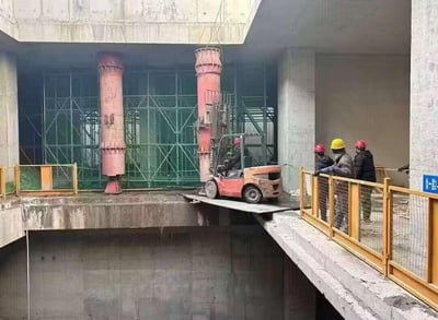 建筑工地施工现场，一辆红色叉车正行驶在临时搭建的金属板桥上，桥面连接着两个施工平台，下方是深基坑。桥的右侧有三名戴着安全帽（黄色、红色）的施工人员，他们正注视着叉车的动向。背景可见绿色脚手架和两根大型红色立柱，整体场景展现了建筑施工中的一个紧张瞬间。