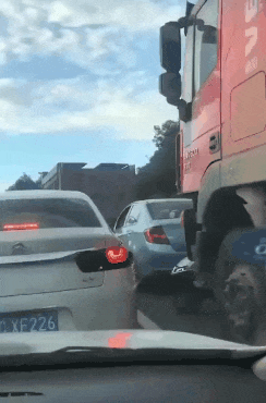 这是一段记录道路堵车场景的动态GIF，画面中狭窄的道路上挤着多辆汽车，其中左侧的白色轿车右侧尾灯被网友P成了愤怒的红色眼睛样式，赋予了车辆拟人化的烦躁情绪，搭配拥堵的路况，生动地调侃了人们在堵车时的郁闷心情，让原本枯燥的堵车场景变得趣味十足。
