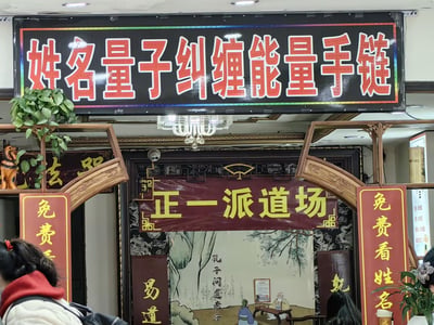 这是一张室内商铺的实拍照片，画面最上方是黑底红字的醒目招牌“姓名量子纠缠能量手链”，将现代量子物理概念与姓名玄学结合；下方是带有中式雕花木质装饰的入口，入口处悬挂红底黄字的“正一派道场”招牌，两侧竖牌标注“免费看姓名”，入口内部的背景墙绘制着“孔子问道老子”的传统场景，还配有“易道”字样。整体呈现出将伪科学概念、道教文化、姓名算命杂糅在一起的奇特商铺风格，主打打着科学旗号的玄学相关商品与服务，没有双关、谐音梗或暗示内容。