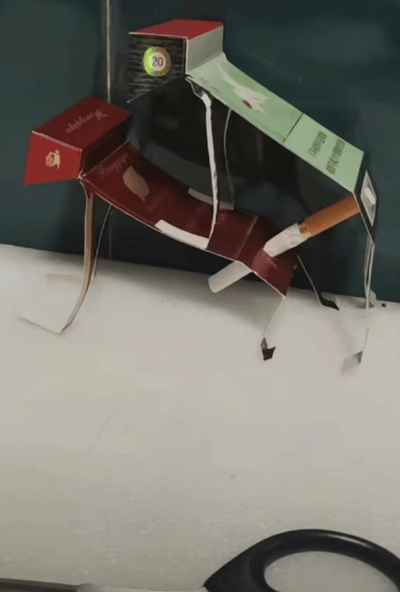 这张图片展示了一个用香烟盒手工制作的小狗雕塑。雕塑主体由多个不同颜色的香烟盒拼接而成，包括红色和绿色的盒子。小狗的头部是一个红色的香烟盒，身体由较大的红色盒子构成，背部还粘贴了一个绿色的盒子作为装饰。雕塑的腿部由切割后的香烟盒纸板支撑，尾巴则是一根真实的香烟。在小狗的背部上方，还有一个带有数字"20"的圆形标识，可能来自香烟盒上的包装元素。整个作品展现了创作者的环保创意和手工技巧，将废弃的香烟盒变成了生动有趣的动物造型。
