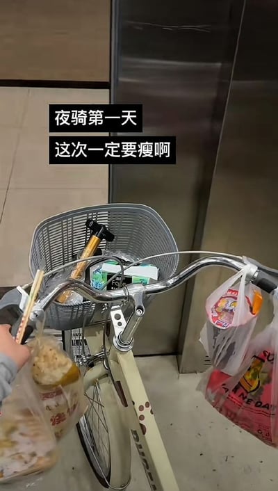 整张图为第一视角实拍，场景位于电梯内，画面主体是一辆米白色带车筐的自行车，车把上挂着多袋打包好的餐食，拍摄者的手也正拿着装有食物的塑料袋，图上方配有两行黑底白字的文字“夜骑第一天 这次一定要瘦啊”，文字展现的减肥预期和实际买了大量食物的画面形成强烈反差，调侃了想要靠夜骑减肥的人刚出发就忍不住购买食物，减肥计划刚开始就失败的搞笑情况，充满生活化的自嘲趣味。