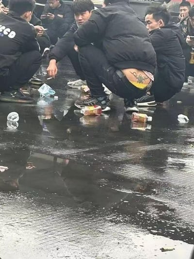 图片中一群人（多为男性）蹲在湿滑的地面上，周围散落着塑料瓶、食品包装盒、烟头和纸巾等垃圾，地面有积水形成的 puddles。画面中央一名男子的裤子明显下滑，露出后腰及臀部位置的纹身图案，纹身颜色鲜艳，图案清晰可见。其他人或低头看手机，或吸烟，姿态随意，整体场景显得较为杂乱邋遢。
