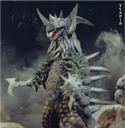 这是特摄剧《泰罗奥特曼》中的暴君怪兽泰兰特的TV版造型截图，怪兽整体造型狰狞凶悍，体表覆盖灰绿与浅灰的怪异纹理，头部生有多根尖角，嘴巴大张露出锋利牙齿，左臂是带刃的巨斧状肢体，右臂附着尖刺流星锤，背部也排列着尖锐突起，背景是烟雾弥漫的昏暗场景，充满了怪兽的压迫感，展现了这只合体怪兽的强大威慑力。