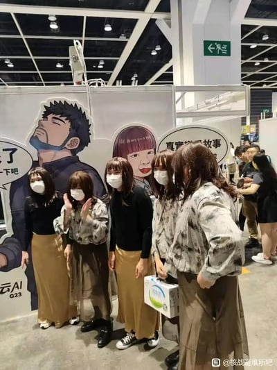 这是在一场漫展的展位前，五位戴着同款假发、口罩的参与者，模仿展位背景板上的女性卡通角色的造型合影。背景板上还有男性卡通角色和带有'真实故事'、'了 一下吧'的对白气泡，以及'云AI G23'的标识。现场还有其他参展的人群，整体氛围轻松欢乐，参与者高度一致的造型让场面充满搞笑趣味。