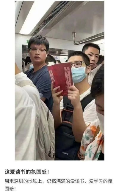 这张图片呈现了周末深圳地铁车厢内的拥挤场景，一位戴着口罩和眼镜的男士正双手捧着一本红色封面、印有“金瓶梅”字样的书籍阅读。周围的乘客或看向镜头或被人群遮挡，图片下方配文称这是“爱读书、爱学习的氛围感”，但由于《金瓶梅》的书籍属性与常规认知里的“学习用书”有差异，形成了一种幽默的反差效果，用调侃的方式记录了地铁里的有趣日常。