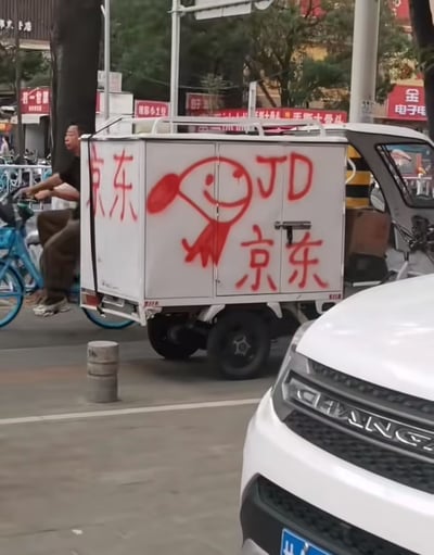 这是一张城市街头的实拍照片，画面主体是一辆停在路边的白色三轮配送车，其车厢外侧用红色喷漆手绘了风格非常随意潦草的京东品牌相关内容：包括歪扭的“京东”汉字、“JD”字母，还有简化卡通版的京东吉祥物Joy小狗的形象，和官方正规的品牌视觉设计形成强烈反差，充满喜感。画面左侧有一名穿黑衣的男子正骑着蓝色共享自行车经过，右下角露出了白色长安汽车的车头局部，背景可见街边的商铺招牌、行道树和交通设施，呈现出普通城市街道的日常景象。