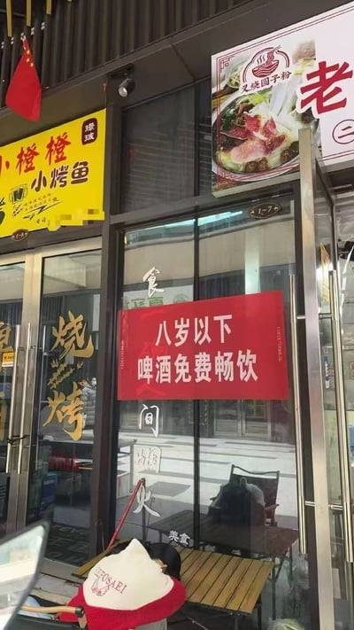 一家餐厅的外立面照片，门口挂着中国国旗。左侧有黄色招牌写着'小橙橙小烤鱼'，右侧有面条的图片和'老'字招牌。最引人注目的是玻璃门上的红色横幅，上面写着'八岁以下 啤酒免费畅饮'，这显然是一个幽默的营销噱头，因为八岁以下儿童不应该饮酒。