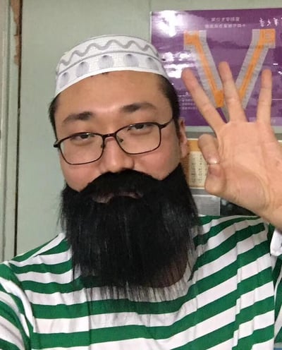 图片中是一位戴着白色花纹帽子、留着黑色胡须的男子，他戴着眼镜，身穿绿白条纹上衣，正对着镜头比出和平手势（V字手势）。背景中可以看到一面浅色墙壁，墙上挂着一张紫色海报，海报上有黄色的V形图案和一些模糊的中文文字。整体氛围轻松随意，像是日常生活中的自拍场景。