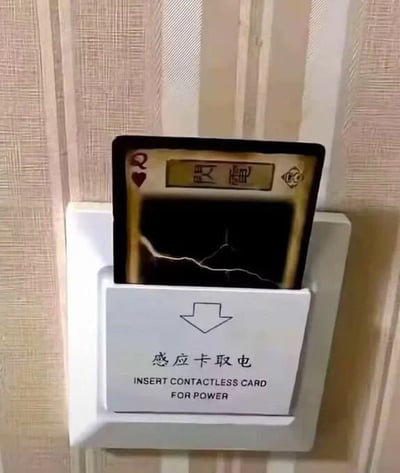 图片展示了酒店房间内的感应卡取电装置，白色面板上印有中文“感应卡取电”和英文“INSERT CONTACTLESS CARD FOR POWER”提示。装置中并未插入常规的酒店房卡，而是被一张红桃Q扑克牌替代，牌面上有金色边框和黑色区域，上方还印有中文“灵魂”二字。整体场景呈现出一种幽默的生活小妙招，用创意方式解决忘带房卡的取电问题，“灵魂”二字增添了趣味性，暗示这张扑克牌仿佛赋予了房间电力。