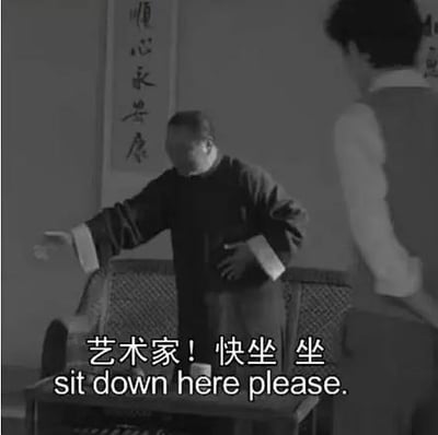 黑白影视截图展现室内场景：左侧穿传统长衫者正伸手热情邀请，右侧穿西式背心者站立旁观。背景可见竖幅书法作品题字“顺心永康”。画面底部叠加对白文字“艺术家！快坐 坐 sit down here please.”，呈现传统与西式元素结合的待客场景。