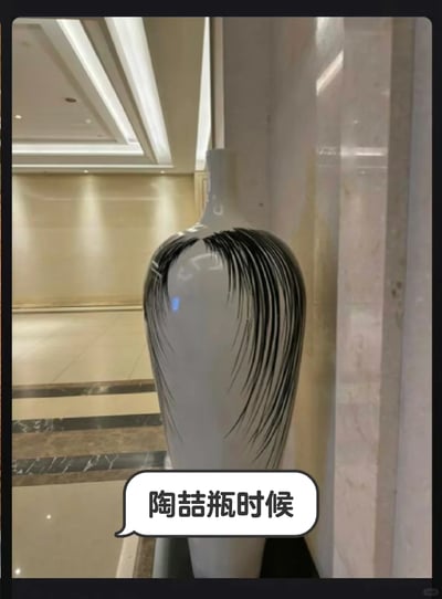 这是一张带有对白气泡的趣味实拍图，拍摄场景为装修精致的室内走廊，地面和墙面采用浅米色石材，顶部有嵌入式灯光设计。画面主体是一个造型修长的浅色花瓶，瓶身的黑色装饰线条呈现出类似歌手陶喆经典刘海发型的视觉效果，下方的对白气泡配有文字“陶喆瓶时候”，玩了中文谐音梗，将“到了瓶颈时候”谐音为“陶喆瓶时候”，结合花瓶的“瓶”与“瓶颈”的关联，再加上瓶身图案和陶喆发型的巧合，制造出幽默搞笑的效果。