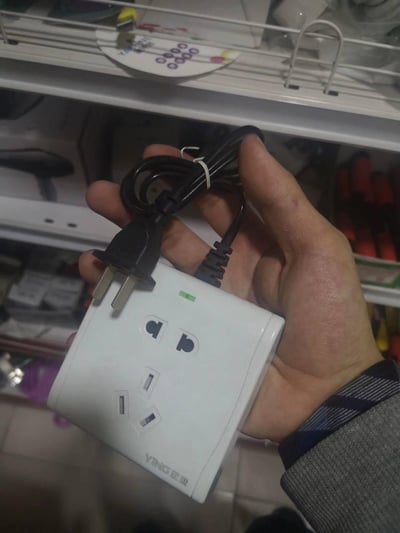 一只手握着一个白色的迎（YING）牌带线电源转换插排，插排上设有一组两孔插座和一组三孔插座，顶部有一个亮起的绿色通电指示灯，插排的电源线与插头被白色扎带捆扎在一起。背景是摆放着各类杂物的货架，整体场景看起来是在店铺内拍摄的商品实拍图。