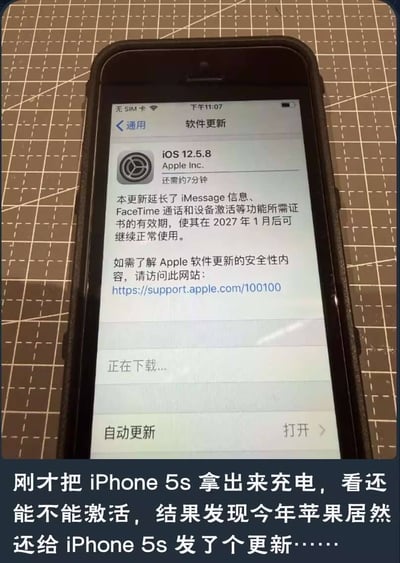 这是一张翻拍手机屏幕的静态照片，主体是装在黑色保护壳里的iPhone 5s手机，放置在带切割格线的切割垫上。手机屏幕显示苹果iOS系统的软件更新页面，正在下载iOS 12.5.8更新，更新说明标注本次更新延长了iMessage信息、FaceTime通话和设备激活等功能证书的有效期，可让该设备在2027年1月后继续正常使用。图片底部配有拍摄者的配文，说明用户翻出停产多年的iPhone 5s充电尝试激活，意外发现苹果仍然为这款老旧机型推送了系统更新，感到十分意外。