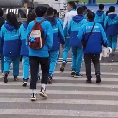 这是一张真实拍摄的静态照片，画面呈现了一群身着蓝白拼色统一校服的学生，正集体走在斑马线上，同行的还有两位成年人：一位背着红白相间的双肩包，包上挂有蓝色小挂饰，穿着深色长裤与运动鞋；另一位背着黑色斜挎包，手中拿着白色物品，穿着深色长裤。背景中可以看到车辆，整体场景大概率是学校组织集体出行或者放学时段的路途中，氛围日常且平和，没有特殊的梗或暗示内容。