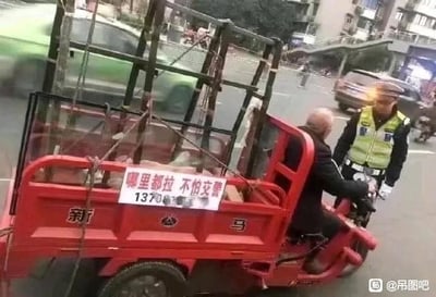 城市街道场景中，一辆红色三轮车停在路边，车斗后部悬挂着白底红字标语牌，上书'哪里都拉 不怕交警'及联系电话。一名身着反光背心的交警正站在三轮车旁，与驾车的老年男子交流。背景可见行驶的绿色出租车、其他车辆及街边建筑，画面因标语内容与实际被交警拦下的情景形成鲜明幽默反差，充满民间调侃意味。