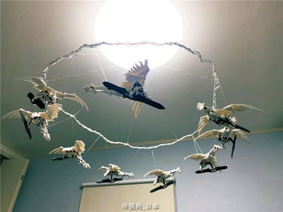 图片展示了一个创意家居装饰场景。天花板中央有一盏明亮的圆形吸顶灯，周围用透明线悬挂着多个带有白色翅膀的机械风格模型人偶。这些模型呈飞行姿态，通过细线连接到一个圆形框架上，整体形成环绕灯光的动态布局。背景是浅色纹理天花板和淡蓝色墙壁，营造出轻盈梦幻的氛围。模型细节精致，翅膀展开，仿佛在空中盘旋。