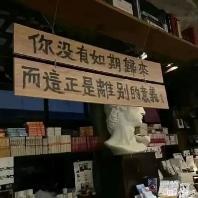 这是一张实拍的静态单图，画面呈现的是一个充满文艺气息的书店或文创店内部场景。画面上方悬挂着两块拼接的木质标牌，标牌上用质朴的毛笔字体写着“你没有如期归来，而这正是离别的意义”，传递出关于离别与遗憾的细腻情绪。标牌下方摆放着一尊西方古典人物的白色石膏半身像，石膏像侧脸朝向画面右侧，神情平静。背景是层层叠叠的书架，上面整齐摆放着各类书籍，整体环境静谧且充满文化氛围，这类陈设常出现在独立文艺书店中，用以营造独特的文艺氛围，吸引文艺爱好者。