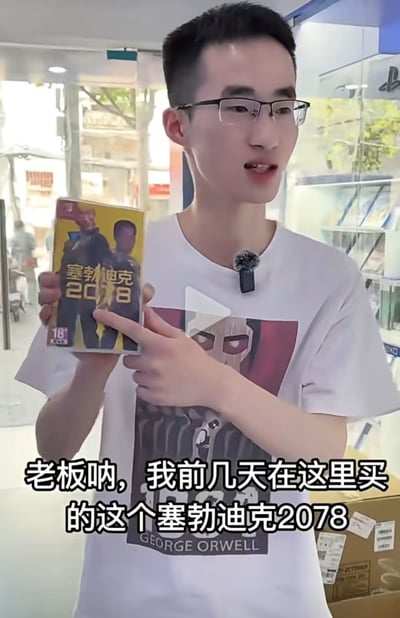 一名戴眼镜的年轻男子在商店内向老板投诉几天前购买的名为《塞勃迪克2078》的游戏。男子手持游戏包装盒，上面印有18+的年龄限制标识。他穿着印有'GEORGE ORWELL'字样的白色T恤，胸前佩戴着麦克风，暗示这可能是一段视频记录或评测内容。商店背景中可以看到玻璃展示柜和疑似PlayStation的标志，表明这是一家游戏或电子产品商店。
