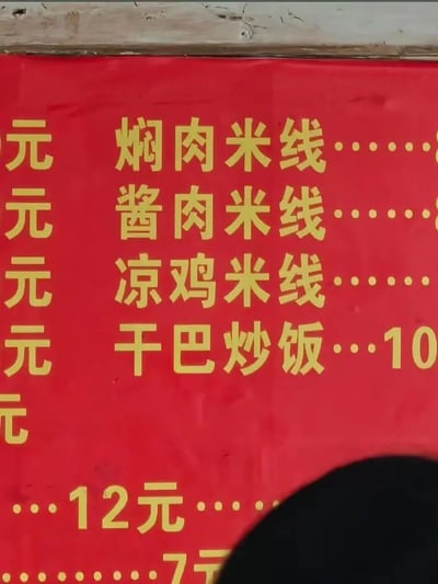 这是一张线下小吃店的价目表局部实拍照片，整体采用红底黄字的醒目配色，可辨认出的在售餐品有焖肉米线、酱肉米线、凉鸡米线、干巴炒饭，其中干巴炒饭标注售价为10元，下方还可见未对应餐品的12元、7元价格标识，价目表最左侧的价格列、上方餐品的价格以及最下方的餐品信息均被截断，右下角存在一块黑色遮挡物，价目表上方边缘能看到粗糙的木质台面或墙面痕迹。