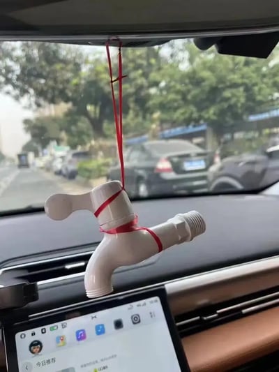 这张图片展示了一辆汽车的后视镜下，吊挂着一个用红绳绑起来的塑料水龙头，前方放着智能手机导航。开车时挂水龙头并不是常规操作，这种不寻常的组合制造出强烈的荒诞和幽默感。这个场景常常被网友玩梗，用来调侃“车上不缺水”、“挂件创意”、“我这儿水多”等暗示梗，常见于社交媒体爆梗图里。