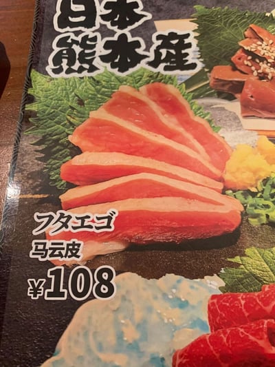 这是一张翻拍的日式料理店纸质菜单页面，页面顶部有日文“日本熊本产”，核心展示的是一款标注为“马云皮”（对应日文“フタエゴ”）的菜品，呈现为几片红白相间的生切水产食材，放置在绿色装饰叶上，搭配有黄色山葵、绿色葱花作为配菜；页面右上角还有另一款撒了白芝麻的深色菜品。该菜品售价为108元，其中“马云皮”是日文食材名的错误音译，实际为日本熊本产的特定水产部位，属于菜单翻译时出现的趣味乌龙，和人物马云并无关联。
