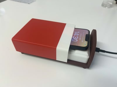 这是一张实拍的趣味数码配件图，展示了一款模仿日式便当盒造型的手机充电底座。红色的盒身搭配白色内托，手机放置在底座中，露出的屏幕部分如同便当里的特殊食材，创意十足。手机屏幕显示时间为14:30，还有日期和电量信息，底座连接着充电线正在为手机供电，兼具实用性与趣味性。