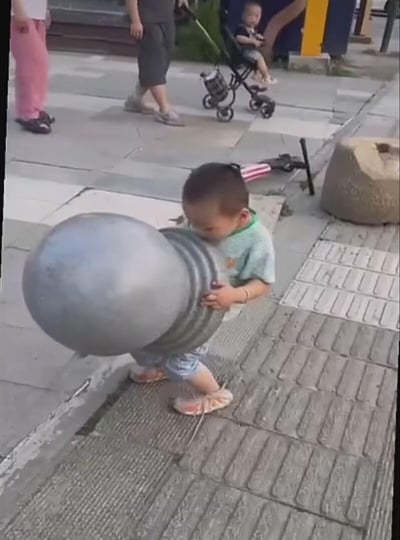 街头抓拍的趣味一幕：一名穿着浅绿色上衣的幼儿正双手抱着一个比他脑袋还大的金属球，努力保持平衡走在人行道上。小家伙表情专注，身体微微前倾，展现出与年龄不符的“大力士”气质。背景中可见其他行人路过，有人推着婴儿车，营造出真实的城市生活氛围。整个画面充满童真与意外的反差萌，让人忍俊不禁。