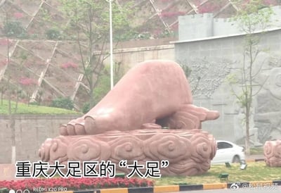 这是一张拍摄于重庆大足区的实拍照片，画面核心是一座巨型的脚形雕塑，雕塑以祥云造型为底座，安置在道路旁的绿化区域中。背景可见带有护坡结构和绿植花卉的坡地，还有一辆白色轿车从雕塑旁的道路驶过。
图中配文点明这是重庆大足区的“大足”，用巨型脚的雕塑来呼应“大足”这个地名，是将地名做了谐音+具象化的趣味设计，充满巧思，让人一眼就能get到和地名的关联。