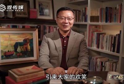 这是一段访谈类视频的截图，画面中一位戴眼镜的男士身着浅灰色条纹西装外套与酒红色内搭，坐在布置有书籍、装饰画的室内场景中，双手置于桌前，面带温和的微笑，似乎正在分享故事或讲述内容。画面底部配有白色字幕“引来大家的欢笑”，左上角带有“观传媒 GUAN MEDIA”的平台水印，右下角标注有“回忆与点”的栏目标识，整体氛围轻松愉悦，给人一种亲切的讲述感。