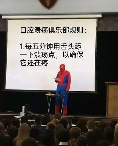 图片展示了一个类似教室或 lecture hall 的场景，舞台中央站着身穿红蓝配色蜘蛛侠 costume 的人，手持纸张站在讲台后，面对台下坐着的众多观众。背景的投影仪屏幕上醒目地显示着“口腔溃疡俱乐部规则：1.每五分钟用舌头舔一下溃疡点，以确保它还在疼”的黑色文字。画面通过蜘蛛侠这一严肃的超级英雄形象与荒诞的“俱乐部规则”形成反差，幽默地调侃了人们患口腔溃疡时，明明知道舔伤口会疼却忍不住反复触碰的常见生活体验。