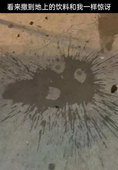 这是一张带有文字加注的原始拍摄静态图，画面中干燥的地面上洒出的深色饮料晕开形成水渍，巧合地构成了一个有着瞪大的双眼、张开的嘴巴的惊讶表情图案。图片上方配有文字“看来撒到地上的饮料和我一样惊讶”，通过拟人化的手法，将偶然形成的水渍图案与人的情绪关联起来，制造出诙谐有趣的效果，记录了生活中一个充满巧合的趣味瞬间。