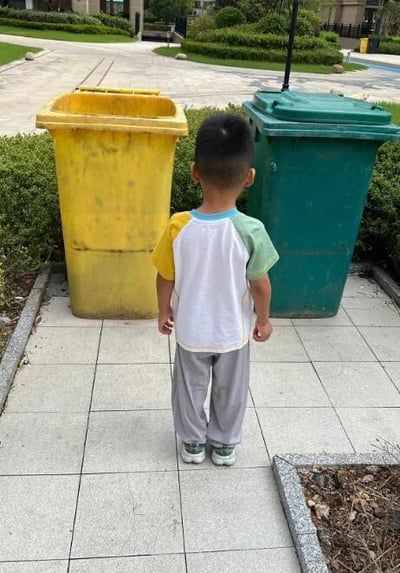 一个小男孩背对着镜头站在两个垃圾桶前，他穿着白色为主、袖子带有黄色和绿色拼接的短袖T恤，搭配灰色长裤和绿色运动鞋。面前是一个黄色垃圾桶和一个绿色垃圾桶，放置在铺有灰色地砖的小路上。背景可见住宅小区的绿化景观，包括修剪整齐的灌木丛、草坪以及远处的居民楼和道路，整体场景呈现出日常社区环境的平静氛围。