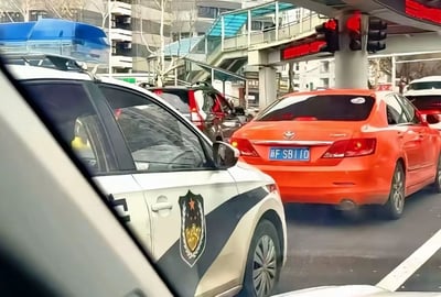 这是一张从车内视角拍摄的城市路口实拍静态图，拍摄时交通信号灯显示红灯，多辆机动车在路口停止线后等待通行。画面左侧是一辆带有警用标识、顶部装有蓝白警灯的制式警车，右侧紧邻的是一辆橙色丰田轿车，该车悬挂的蓝色机动车号牌为浙F·SB110，号牌字符组合本身带有戏谑调侃的含义，又恰好出现在警车旁，形成了充满巧合的滑稽效果。画面背景可见带玻璃顶棚的人行过街天桥、临街居民楼与光秃的行道树，是典型的北方城市日常街景。