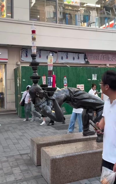 城市商业街区的户外场景，画面中央是一座黑色金属材质的抽象人物雕像，雕像呈现出身体前倾、单手支撑的动态姿势。有趣的是，雕像的手掌上被人创意性地堆叠了多个不同种类的瓶罐，包括饮料瓶、酱料瓶等，形成了一个不稳定但又奇妙平衡的塔状结构。背景中可以看到服装店的橱窗和绿色的施工围挡，有几位行人正从雕像旁经过，整个场景展现了城市公共空间中艺术与日常物品的有趣结合。