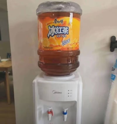 这是一张室内实拍的静态照片，画面主体为一台白色美的牌立式饮水机，饮水机上方没有放置常规的饮用纯净水水桶，而是放置了一款尺寸和饮水机桶一致的定制款康师傅冰红茶桶，桶身印刷的包装设计和普通小瓶装康师傅冰红茶完全相同，桶内盛满了棕褐色的冰红茶液体，可直接通过饮水机的冷热水龙头接取。画面左侧背景处能隐约看到木质餐桌的一角，右侧墙边能看到挂钩和袋装物品的局部，整体场景偏向居家或办公环境。