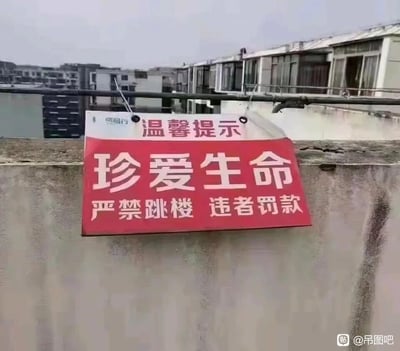 这张图片拍摄于居民楼楼顶，画面主体是一块悬挂在水泥墙上方的提示标语牌。标语牌分为上下两部分，上方白色区域写有“温馨提示”字样，下方红底白字区域写着“珍爱生命 严禁跳楼 违者罚款”。该标语存在明显的逻辑漏洞：若有人真的实施跳楼行为，当事人将失去生命，根本无法执行“违者罚款”的处罚，这种不合理的表述让原本严肃的安全提示变得荒谬可笑，成为了图片的核心笑点。背景中可以看到周围的居民楼建筑，整体场景是普通的城市楼顶环境。