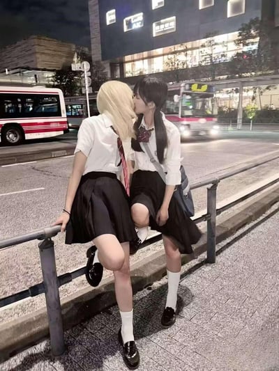 夜晚的城市街头，两位身着JK制服的女生倚靠在马路护栏旁，黑发女生亲吻金发女生的脸颊，背景是行驶的日式公交与城市建筑，整体氛围浪漫温馨，充满青春感，是很有氛围感的百合向日常摄影作品。